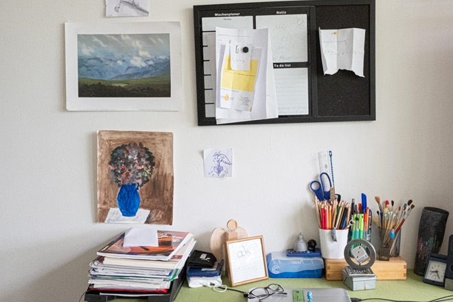 Schreibtisch mit Blätterstapeln und Stiften, im Hintergrund eine Pinnwand und Bilder an der Wand