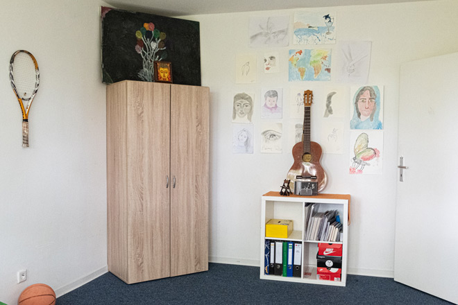 Ein Jugendzimmer mit Kleiderschrank, Sideboard, Gitarre und selbst gemalten Bilder an der Wand.