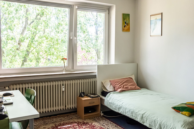 Schlafzimmer mit großem Fenster, Einzelbett und orientalischem Teppich.