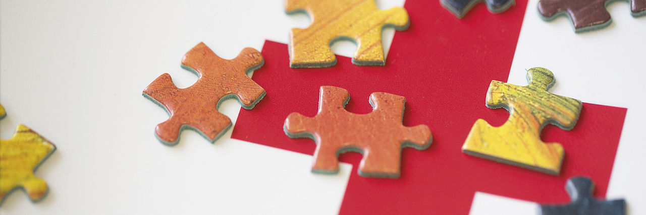 Rotes Kreuz auf weißem Grund, bedeckt mit bunten Puzzle-Teilen.