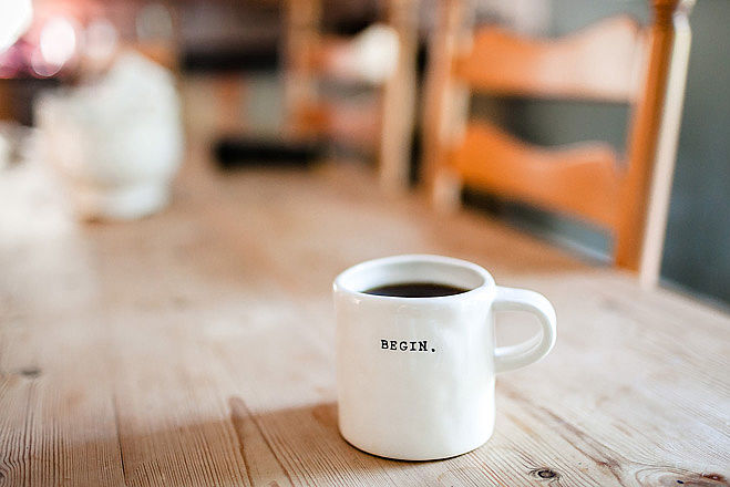 Weiße Kaffeetasse mit dem schwarzen Schriftzug "BEGIN."auf einem Holztisch.