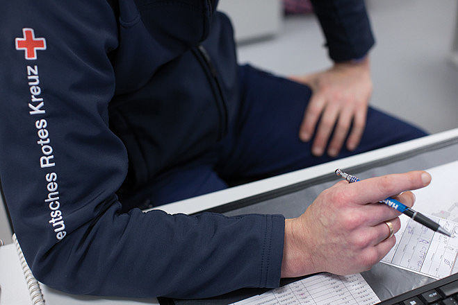 Ein Mann in blauer DRK-Dienstkleidung sitzt am Schreibtisch und hält einen Stift in der Hand.